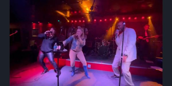 Three energetic singers perform on stage with colorful lights and live band.