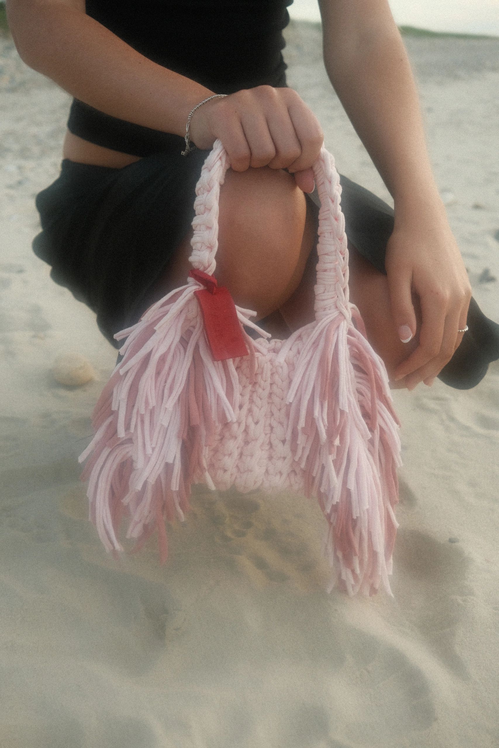 Person holding a pink fringed bag while sitting on the sand.