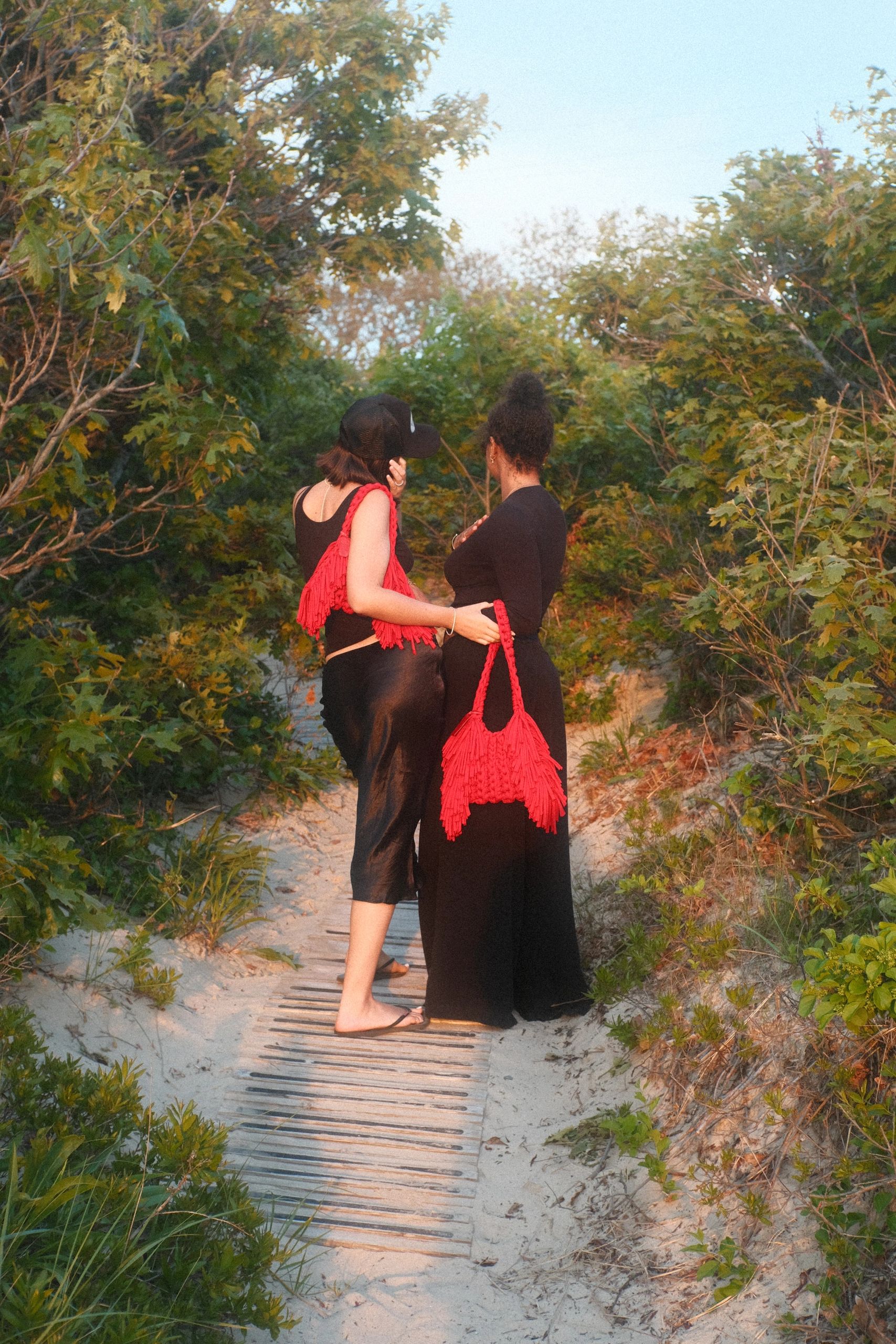 Two women in black dresses with red bags walk on a sandy path surrounded by greenery.