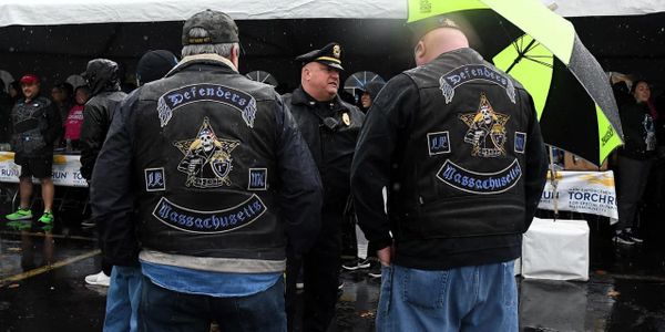 Two men in leather jackets with 'Defenders Massachusetts' patches at a rainy event.