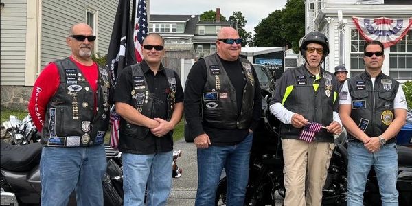Five bikers in leather vests and sunglasses standing in front of motorcycles.