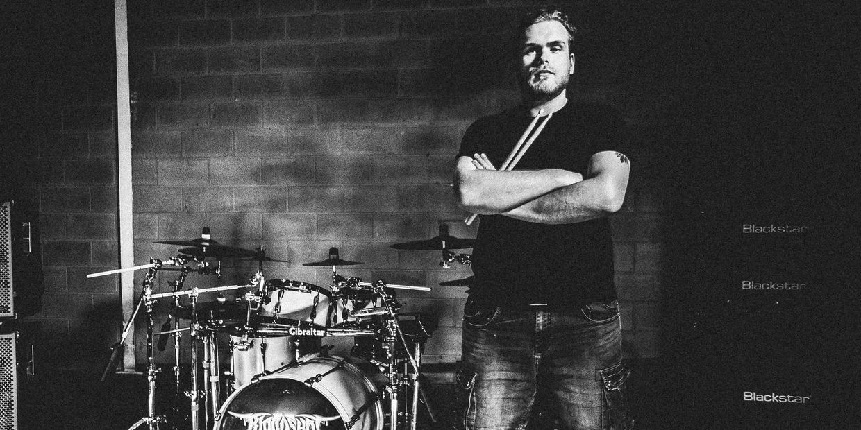 Man standing confidently with drumsticks by a drum set.