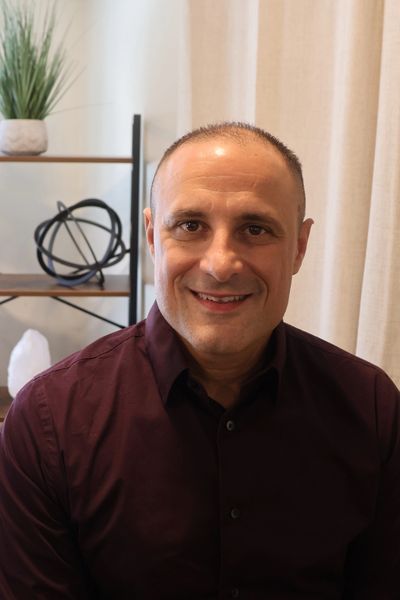 Smiling man in a maroon shirt sitting indoors with home decor in the background.