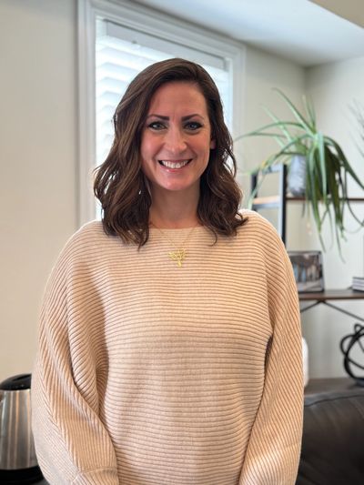 Smiling woman in a beige ribbed sweater standing indoors with a plant in the background.
