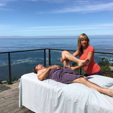 A woman gives a leg massage to a man on a massage table by the ocean.