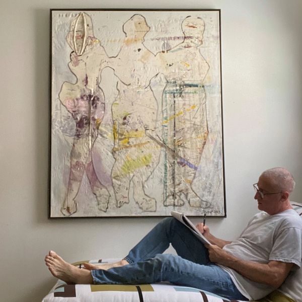 A photo of Keith Theriot lounging on a chair sketching on a notebook next to one of his paintings