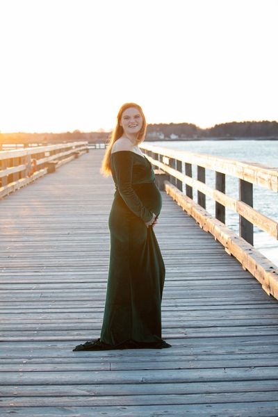 Pregnant woman in a green dress standing on a pier at sunset.