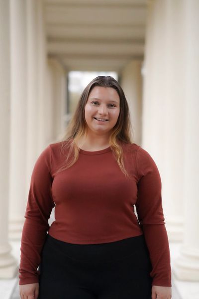 Smiling woman in a rust-colored long sleeve shirt standing in an architectural corridor.