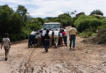 A group of people push a stuck truck on a dirt road.