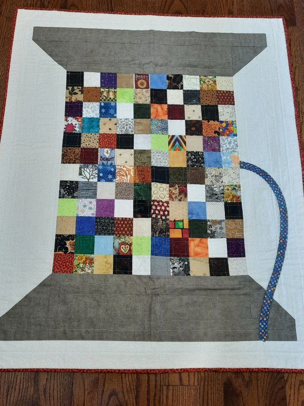 Quilt resembling a colorful thread spool with patchwork squares and a fabric thread.
