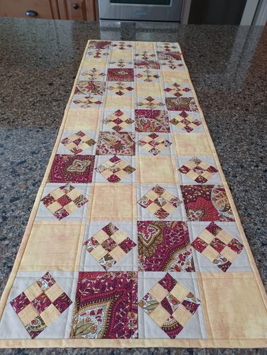 A quilted table runner with beige and red patterned squares on a granite countertop.