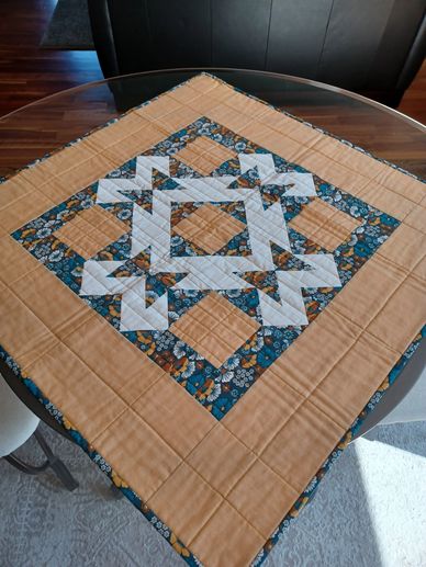 Handmade quilt with geometric patterns in beige, blue, and white on a glass table.