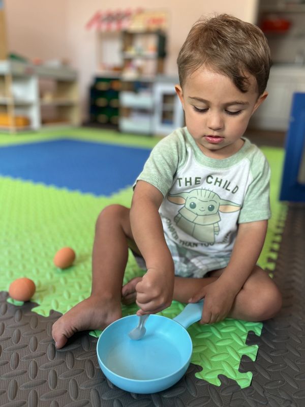 A young child stirs something in a blue pan while sitting on a colorful foam mat.