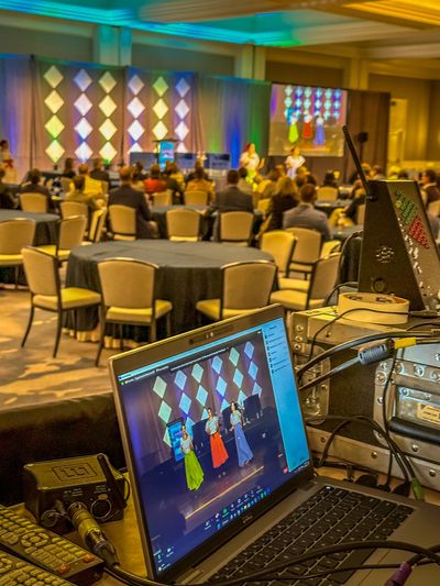 A conference set up as seen from the AV table. Round tables, a stage with decorations and 2 screens 
