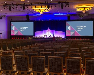 a plenary session with theatre set up, 2 screens saying Technical Summit 2025 and a stage. no attend