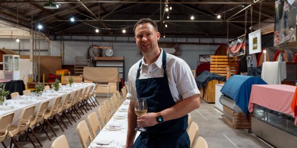 Man in apron holding a wine glass in a rustic dining setup.