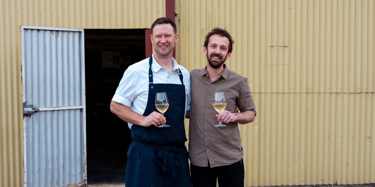 Two men with wine glasses outside a corrugated building.