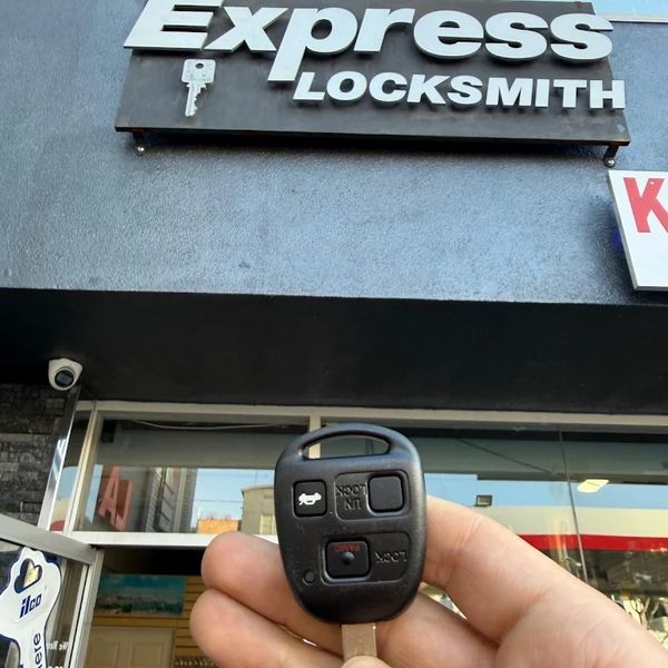 Hand holding a car key fob outside Express Locksmith shop.