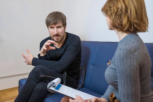 A man with a beard gestures while talking to a therapist taking notes on a clipboard.