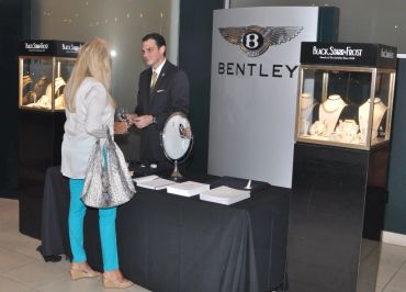 A man in a suit talks to a woman in turquoise pants at a Bentley jewelry display.