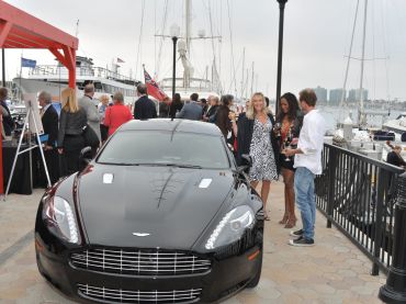 Black luxury car parked at a waterfront event with people socializing nearby.
