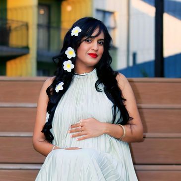 Pregnant woman in a white dress with flowers in her hair, sitting on steps outdoors.