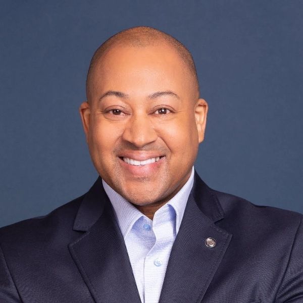 Smiling man in a dark suit jacket and light blue shirt against a blue background.