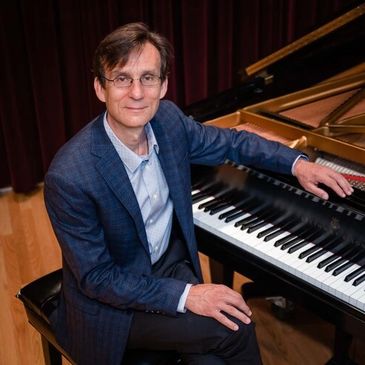 Man in glasses sitting at a grand piano in a formal setting.