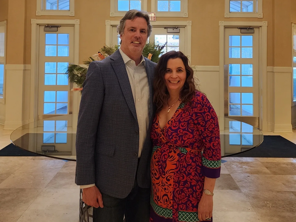 A smiling couple posing indoors in front of large windows and a floral arrangement.