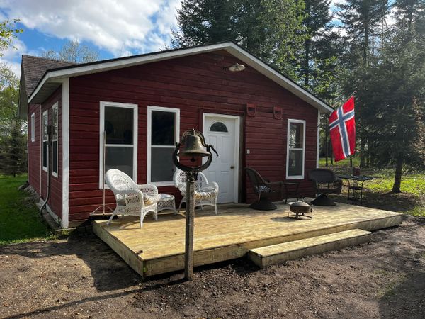 Red cabin with a wooden deck and Norwegian flag in a forest setting.
