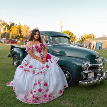 Young woman in a floral quinceañera dress posing by a vintage green truck.