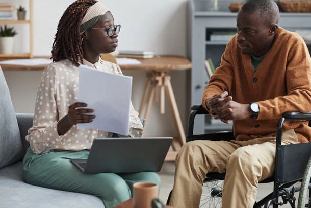 A woman consulting a man in a wheelchair with documents and laptop.