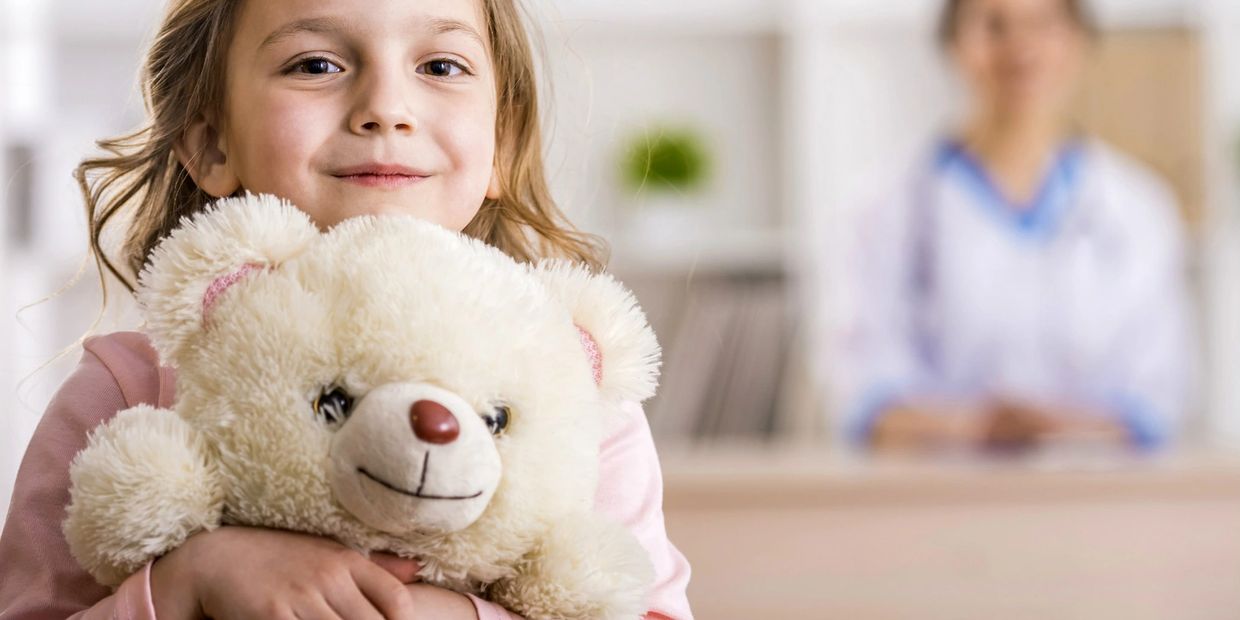Young girl hugging a teddy bear with a doctor in the background.