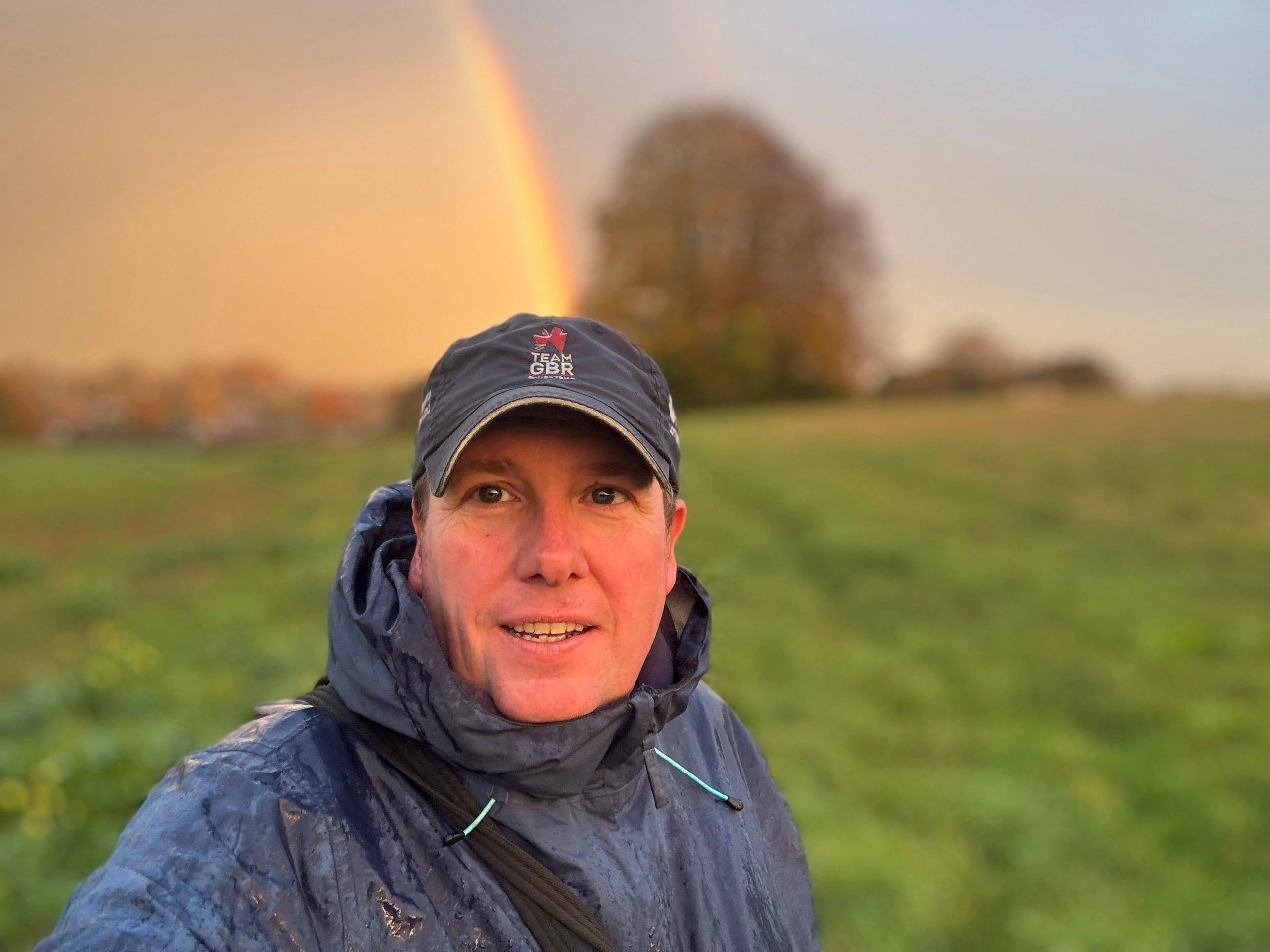 Steve smiling in a rain jacket and cap in a green field, with a rainbow behind him