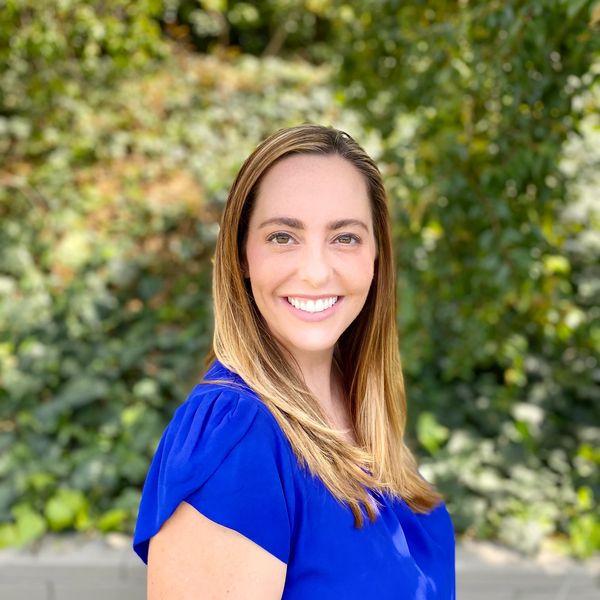 Smiling woman in blue blouse outdoors with greenery background.