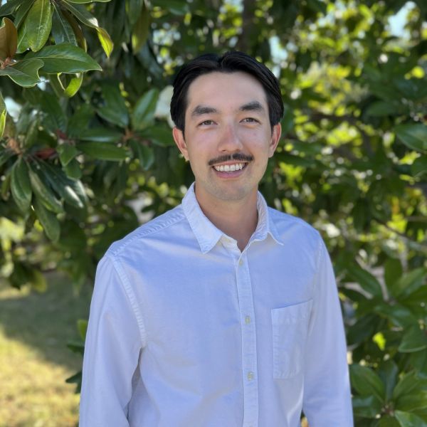 Smiling man in white shirt standing outdoors with green foliage.