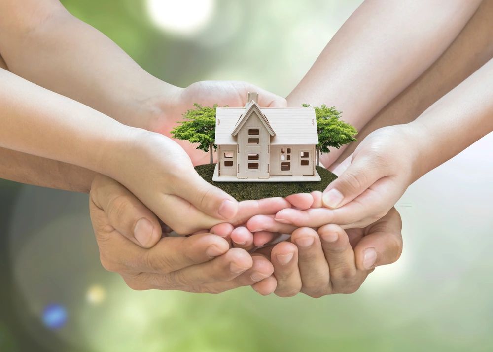 A multitude of hands holding a model of a house with greenery.