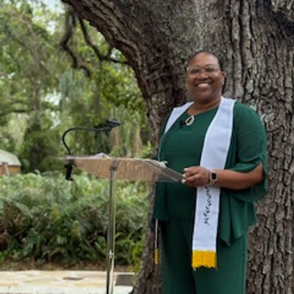 Smiling woman in green outfit speaking outdoors by a tree.