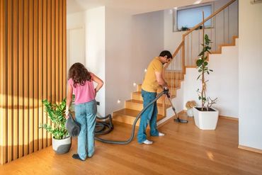 Two people vacuuming wooden floor.
