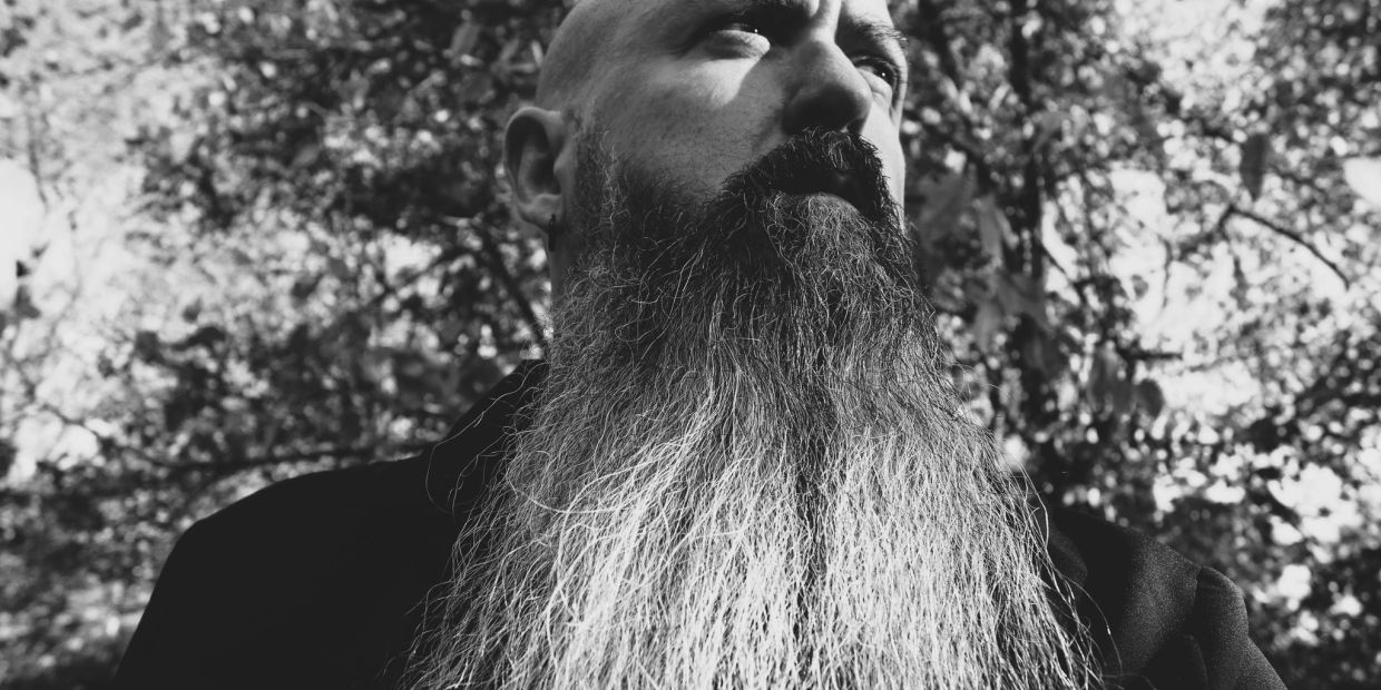 Black and white close-up of a man with a long beard outdoors.