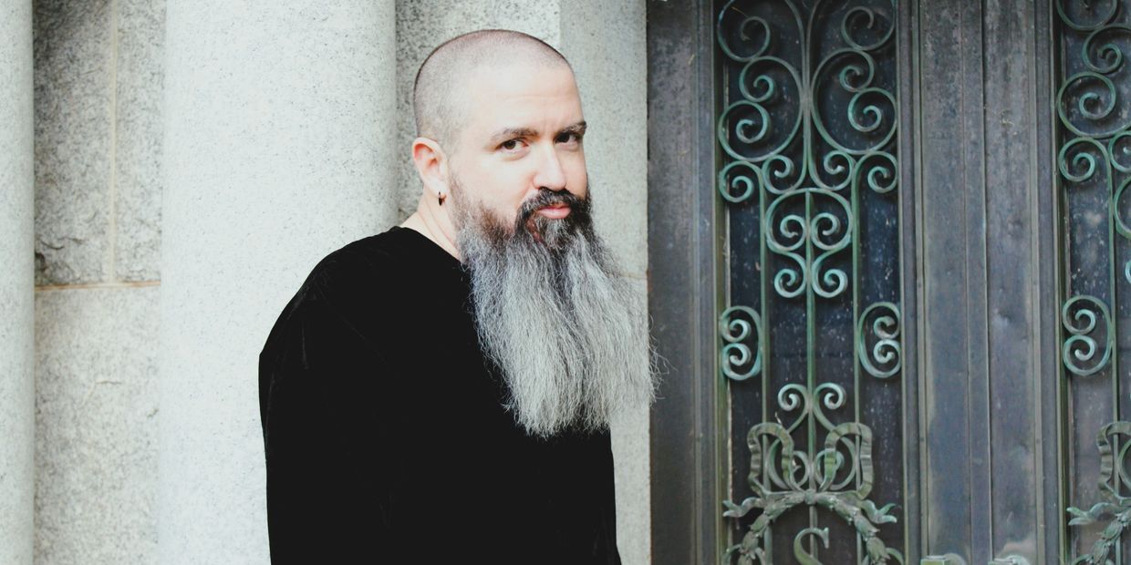 Man with a long beard and shaved head stands by ornate doors.