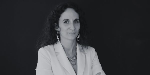 Femme en veste blanche souriante avec cheveux bouclés devant un fond noir.