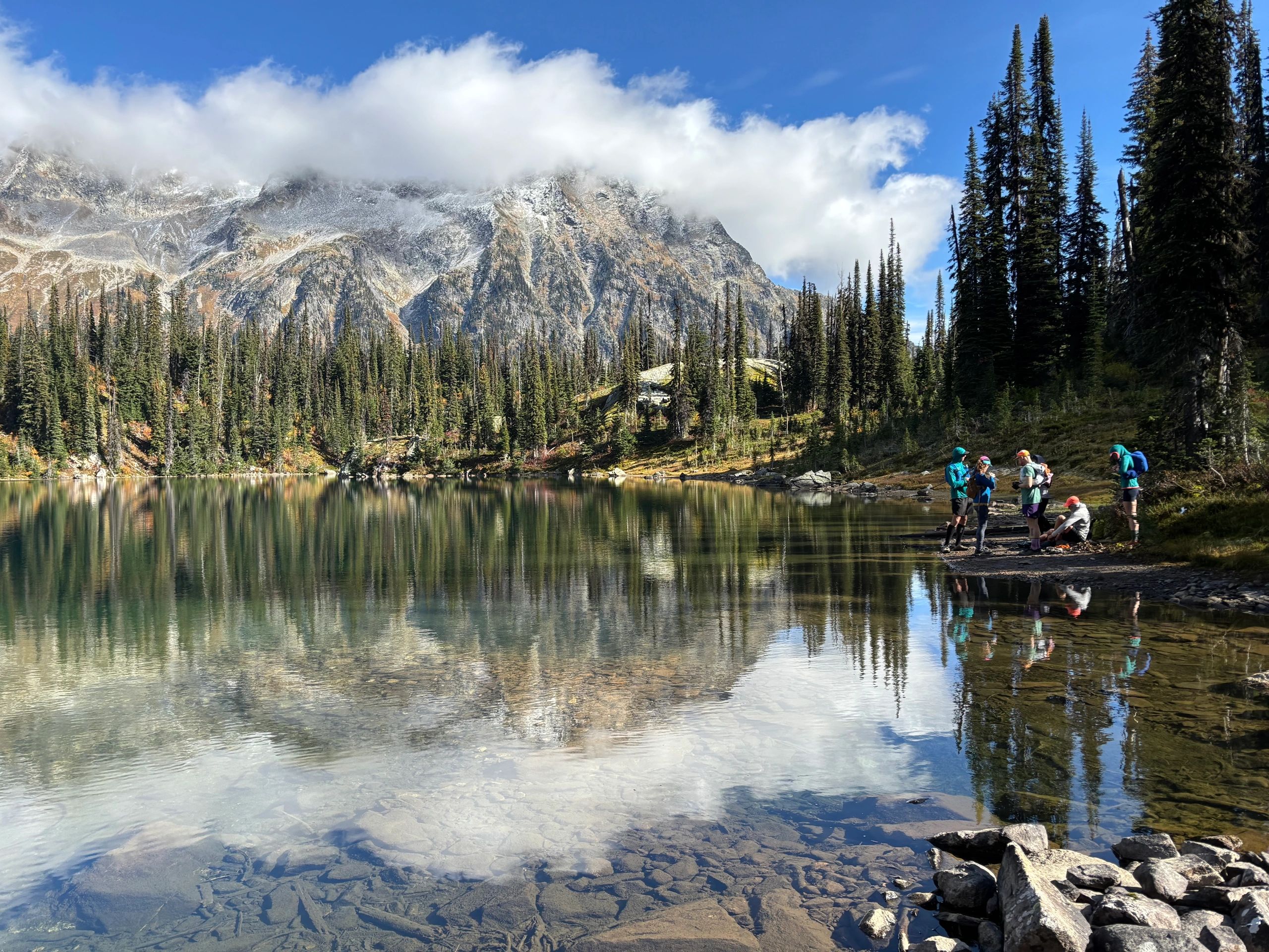 Hikers resting by a serene mountain lake with pine trees and cloudy peaks.