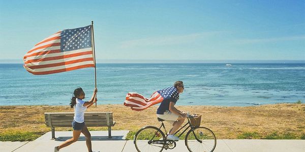 man in bike with American flag draped in  man's back followed by a woman running with American flag