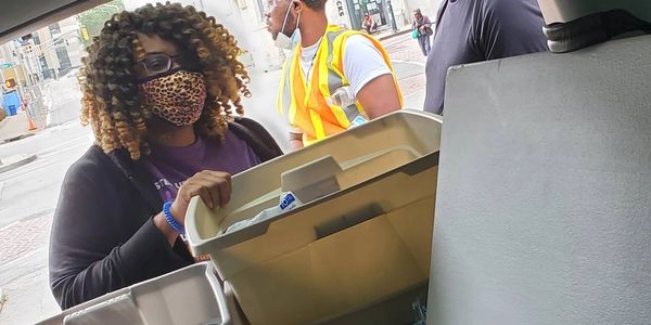 People wearing masks and safety gear handling large plastic bins outdoors.
