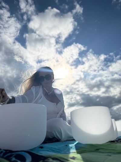 Woman in white with sunglasses, sitting by white bowls under a cloudy sky with sunlight.