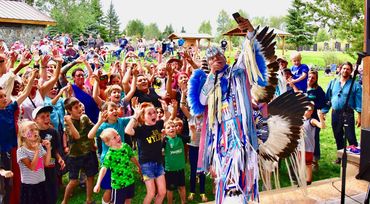 A Native American dancer performs in front of an excited crowd of children and adults outdoors.