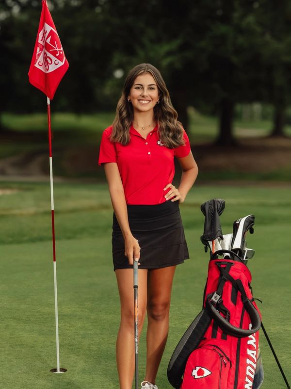 Young woman golfer in red shirt and black skirt standing on green with golf bag.