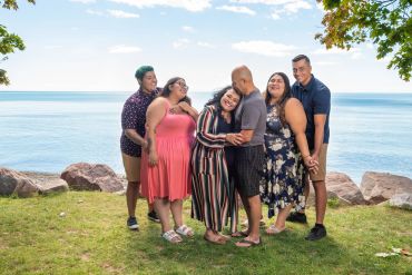 Six people posing happily by a lakeside on a sunny day.