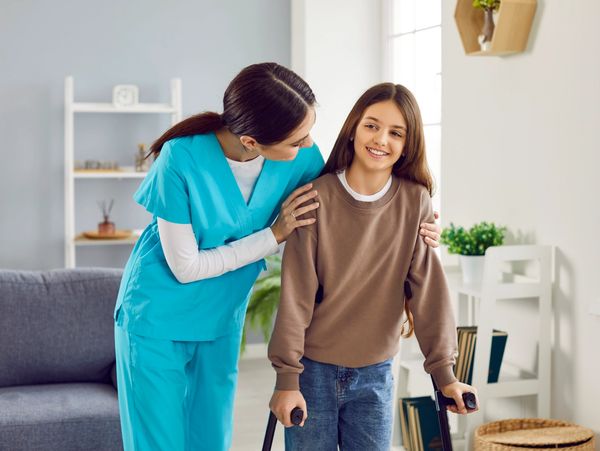 A home healthcare nurse assisting a smiling young girl using crutches.